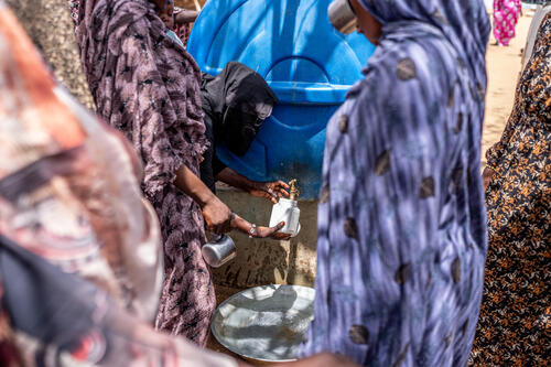 A women pours water into a cup from a storage tank. The water supply in Al Salam Camp is currently considered highly critical and unstable. Access to clean drinking water is insufficient for many families. The poor water supply poses several risks, including the spread of waterborne diseases such as cholera and diarrhea, difficult hygienic conditions, and additional risks for malnourished and sick people.