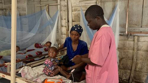 A nurse is checking the pulse and temperature of Faida, a one-year-old hospitalized at the General Referral Hospital of Walikale, supported by MSF, for acute malnutrition, while her mother holds her.