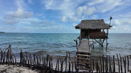 A beach buia pictured in Tebunginako village, Abaiang. This raised structure, often used to live and sleep in, is built from coconut tree wood with a palm-thatched roof offers shade and ventilation from the island heat and is a common sight in Kiribati.  

Kiribati (pronounced Ke-ree-bas) is a vast but climate-vulnerable island nation made up of 32 atolls and one raised coral island in the Pacific. It lies half-way between Hawaii and Australia. With a total land mass of just 811 square km spread over 3.5 million square km of ocean, the country has unique social and geographical challenges. The increase in frequency and intensity of storms and other extreme weather events, the intrusion of salt water with a rising sea, along with declining rainfalls and higher temperatures, threaten ecosystems and are rapidly deplete freshwater supplies.