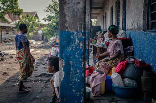 
Des femmes, dont certaines sont victimes de violences sexuelles, et leurs enfants, sont assis à l’extérieur d’une école avec leurs matelas, vêtements et autres effets personnels. Ces familles se sont réfugiées dans l’établissement scolaire après avoir fui les violences. Elles doivent sortir leurs affaires chaque matin pour laisser la place aux élèves pendant les cours. Leurs maris, militaires, ont disparu lors de l’offensive des M23 sur la ville de Goma en janvier et février 2025. 

Women, some of whom are sexual violence survivors, sit with their children outside a school in Goma. These sought refuge at the site after fleeing violence following the M23 offensive in early 2025. Their husbands, soldiers, have disappeared. Each morning, they must move their belongings, including matresses, clothes and other personal belongings, out of the classroom to make room for students.